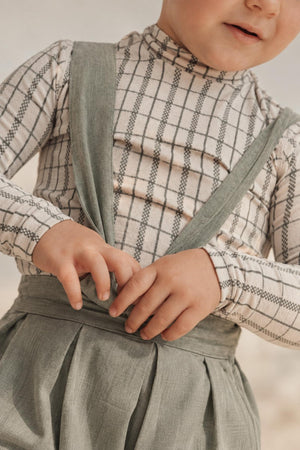 A child is wearing a sage check printed long sleeve cotton turtleneck paired with a stylish, light-colored suspender outfit, showcasing a cozy and playful look perfect for kids.