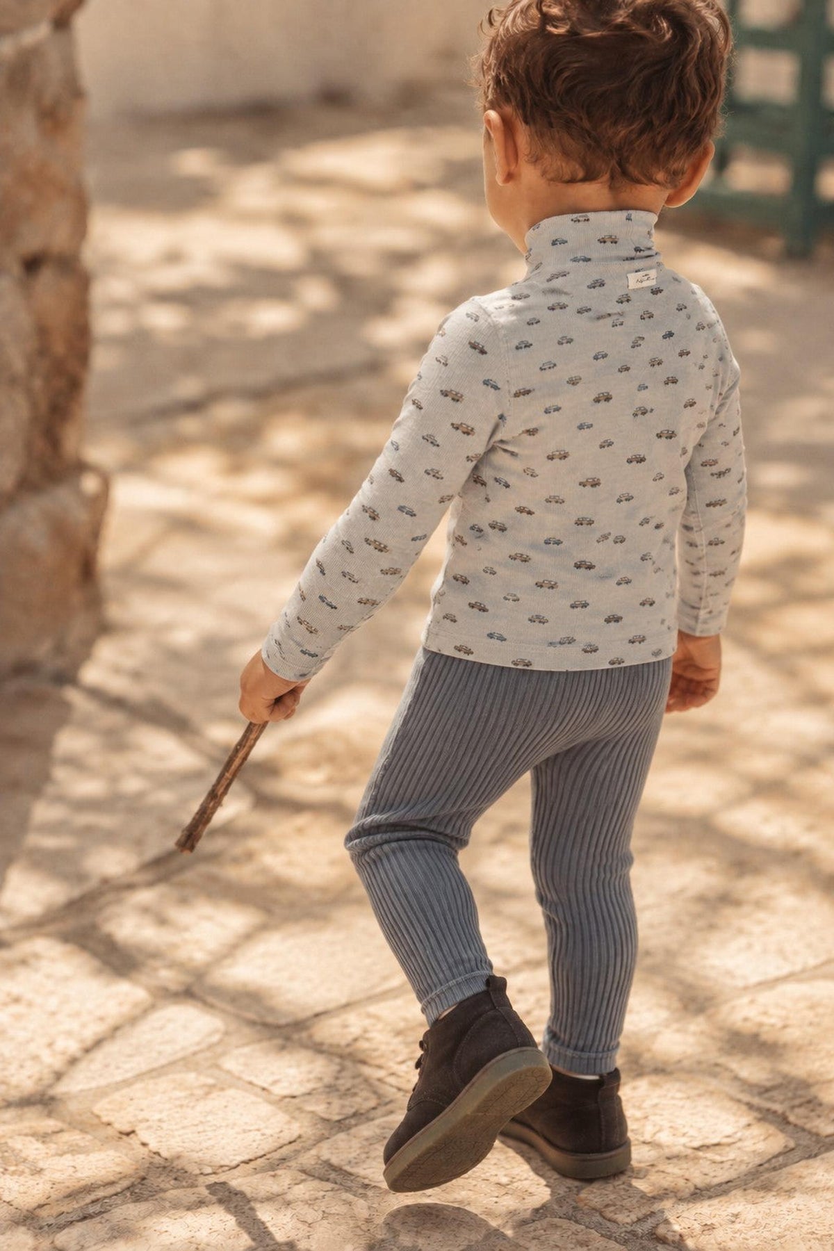 A toddler walks outdoors in a light blue patterned top and ribbed slate cotton leggings, holding a stick. The ground is stone, and soft sunlight casts a warm glow.