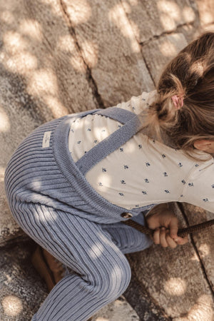 A toddler wears a soft slate blue knit overall paired with a cream shirt featuring a delicate print.