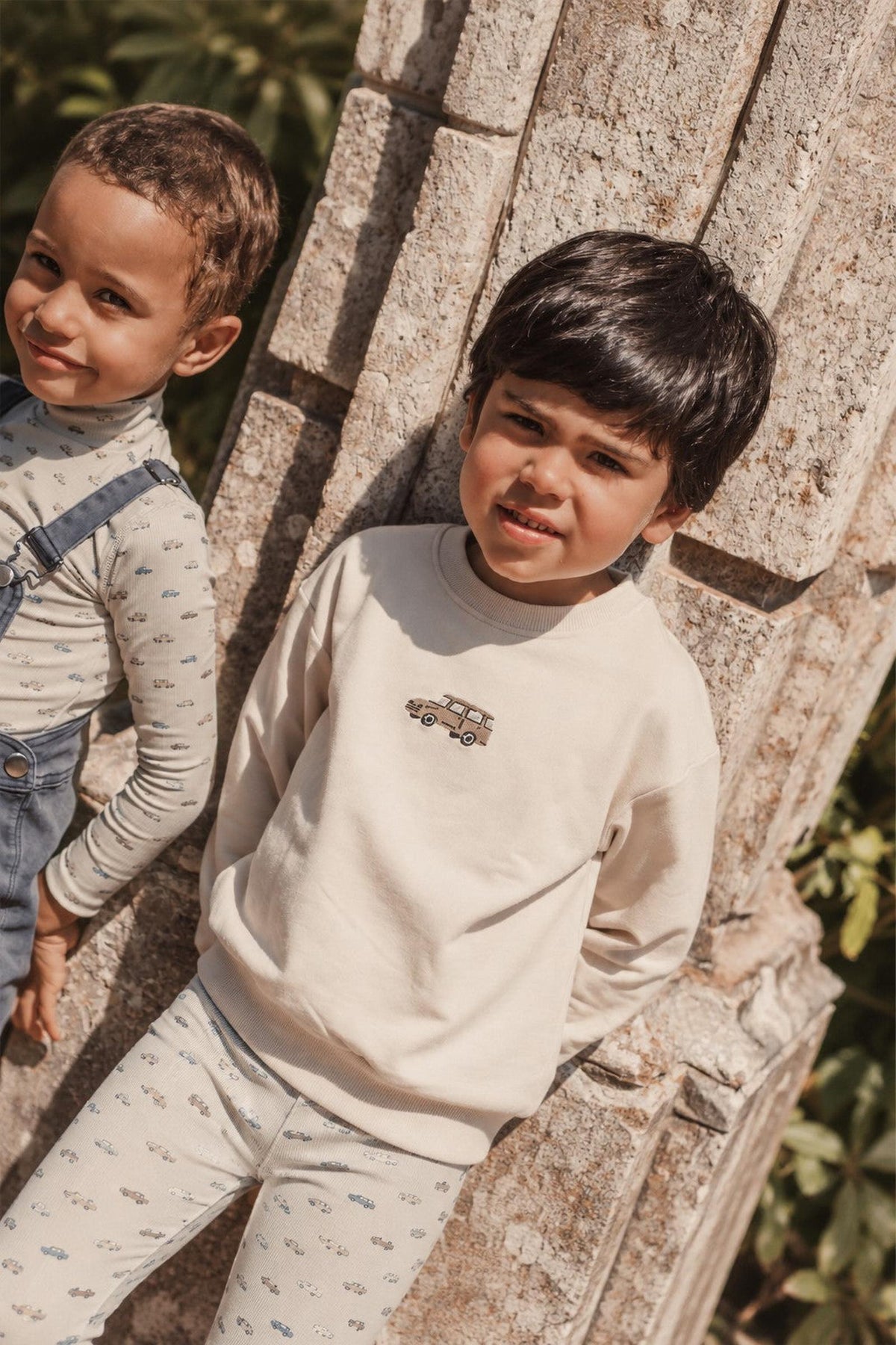 Two young boys are posing outdoors, one wearing a light beige sweatshirt with a vintage car embroidery and the other in a striped long-sleeve shirt and denim overalls.