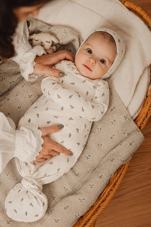 A baby wrapped snugly in a white cotton gown with a leafy pattern, lying in a woven basket. The baby wears a matching hat and looks curiously at the camera.
