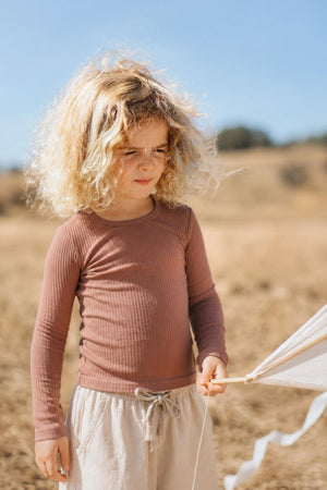A young child with curly hair wears a rosewood ribbed long sleeve tee, holding a kite in a sunny, outdoor setting with dry grass and hills in the background.