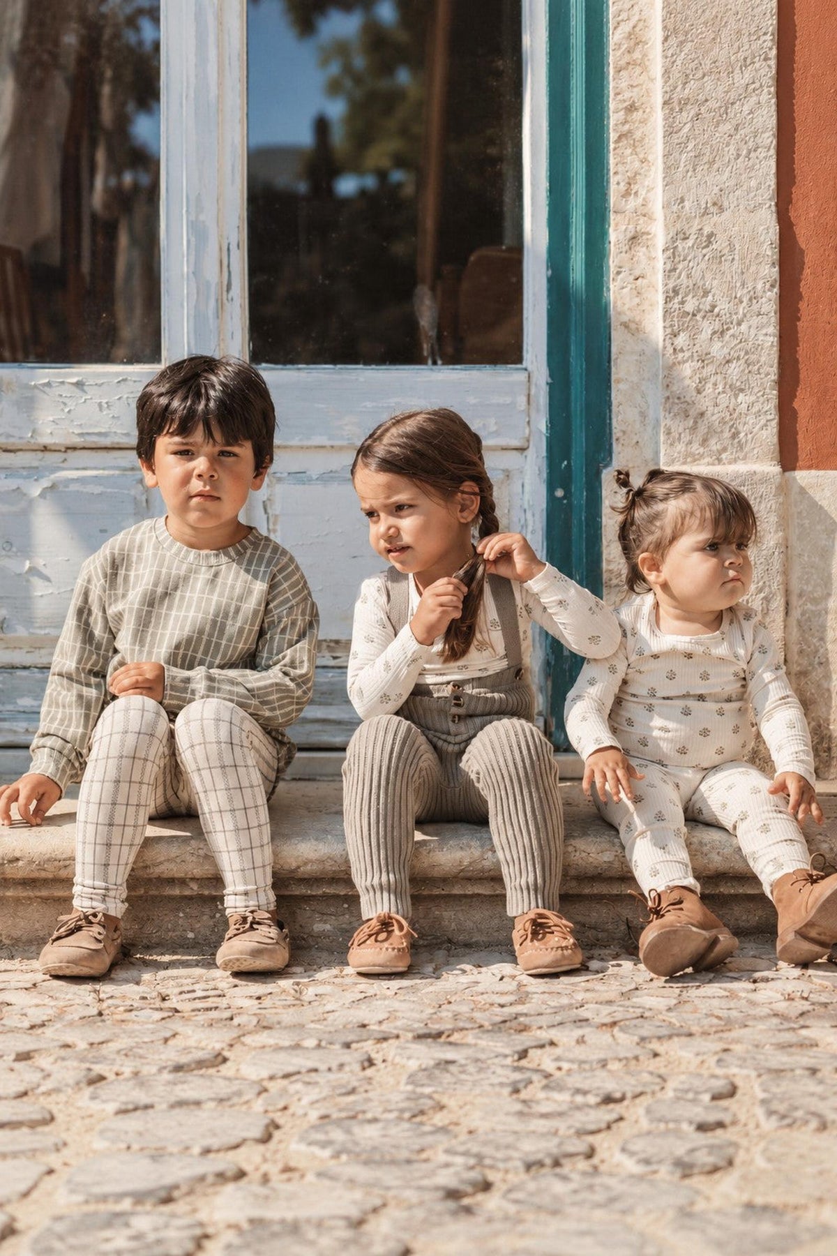 Three children sit on a cobblestone surface, wearing cozy cotton outfits in neutral tones. The scene captures a playful moment outside a rustic building with blue-shuttered windows.