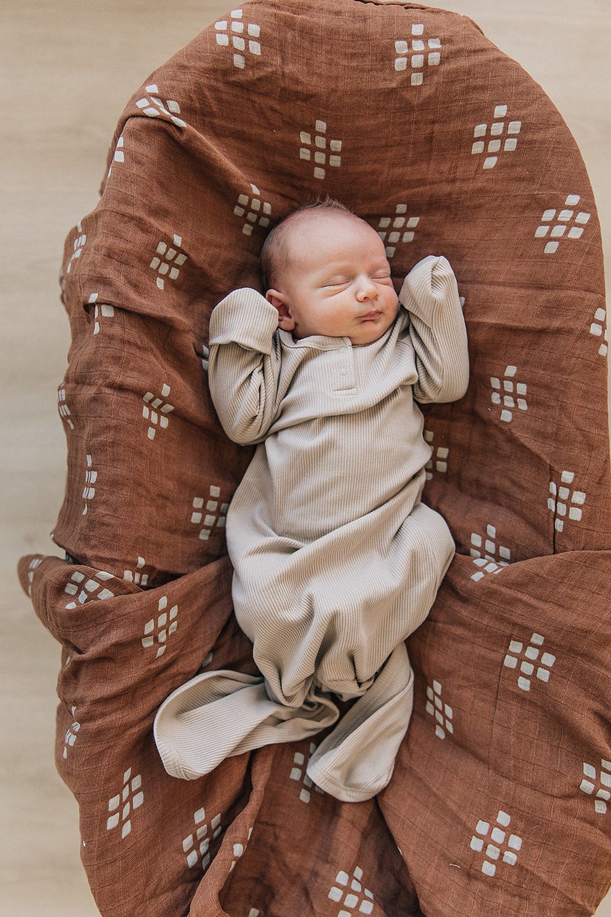 A newborn sleeps in a beige ribbed knot gown on a brown patterned blanket, creating a cozy setting.