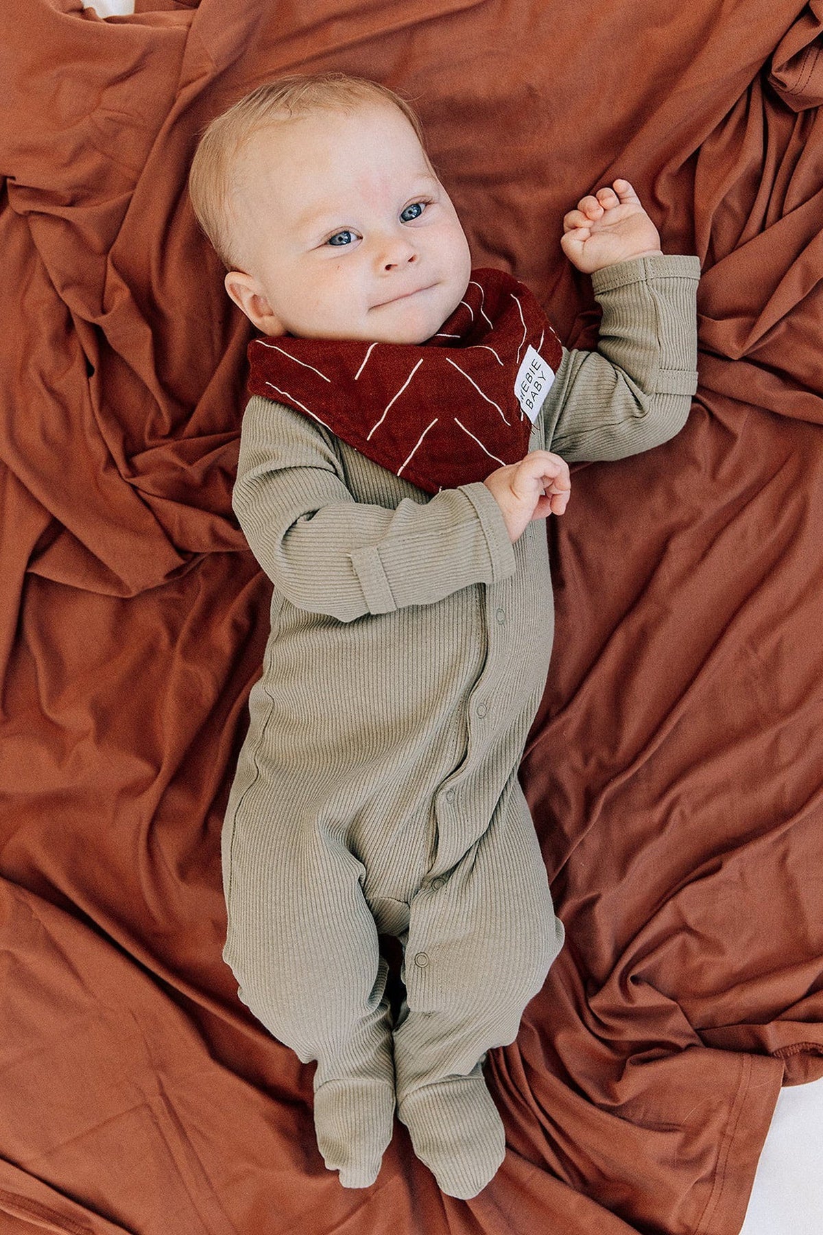 A baby wearing a rust-colored Mebie Baby bib and green ribbed onesie lies on a rust-colored fabric.