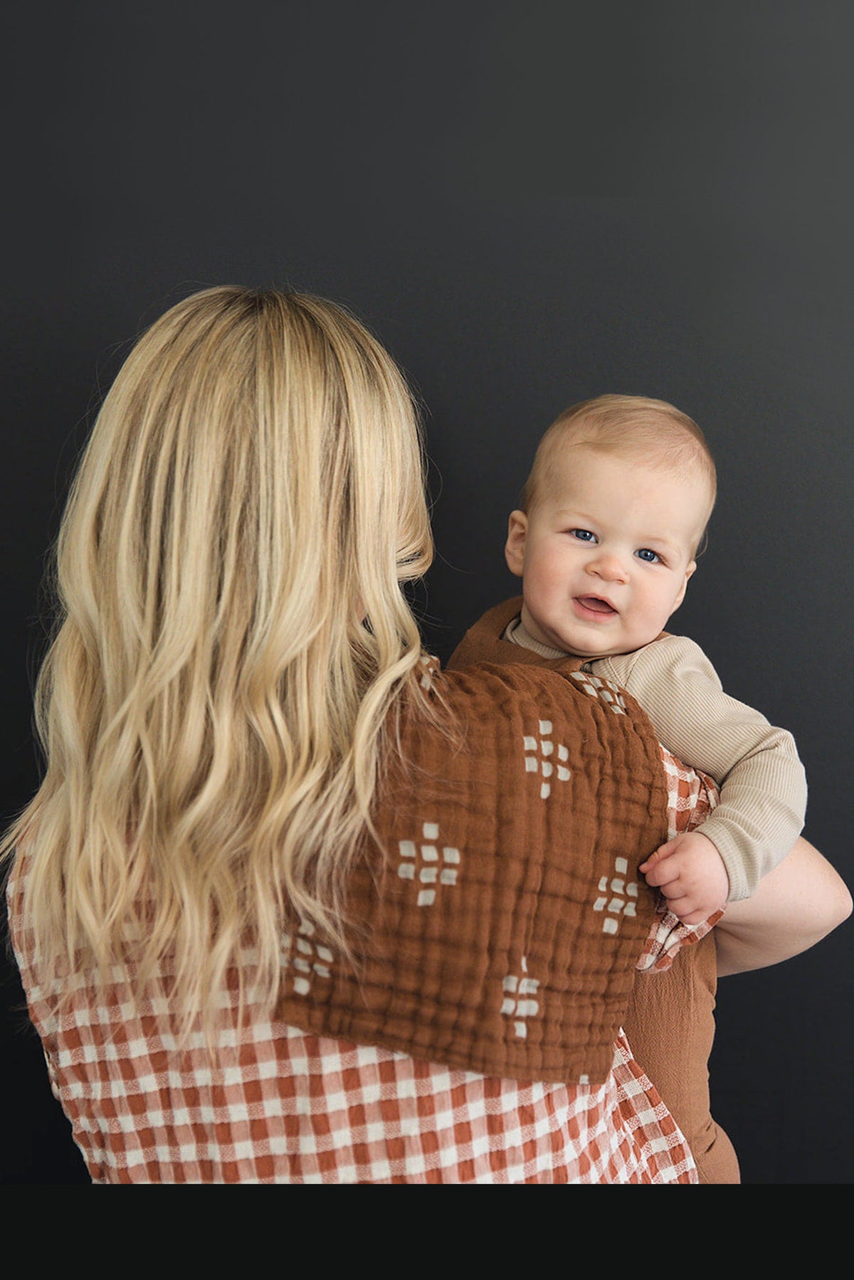 A baby is held by a woman wearing a chestnut muslin burp cloth with a checkered pattern.