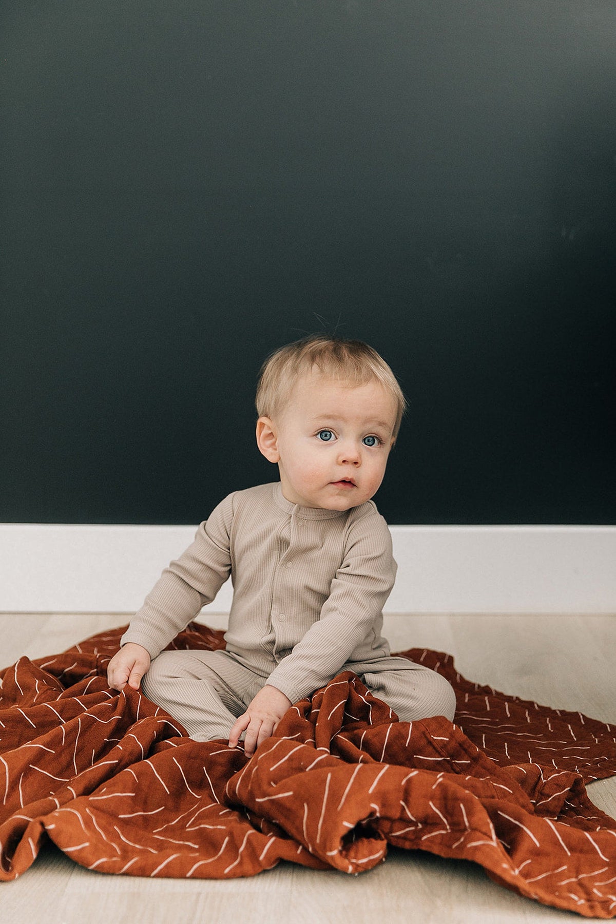 A baby wearing oatmeal-colored ribbed pajamas sits on a rust-colored blanket with white patterns.