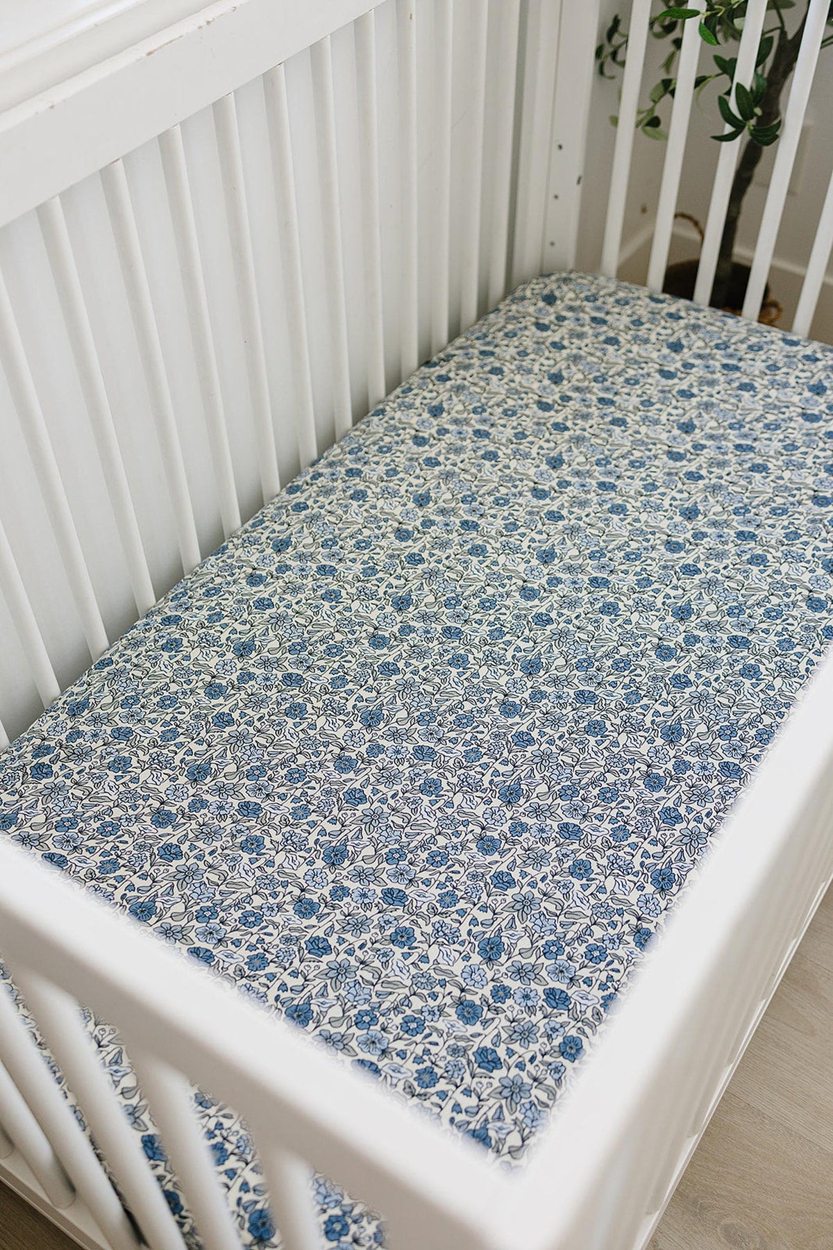White crib with a floral-patterned crib sheet in blue and white. The crib is set in a light, airy room.