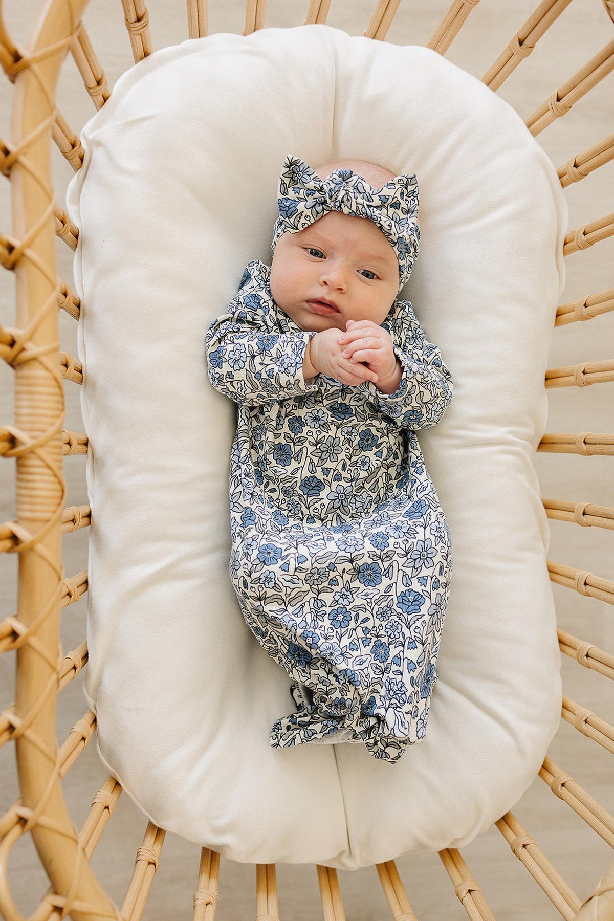 A baby lies in a white cushioned basket, wearing a blue floral gown and matching headband.