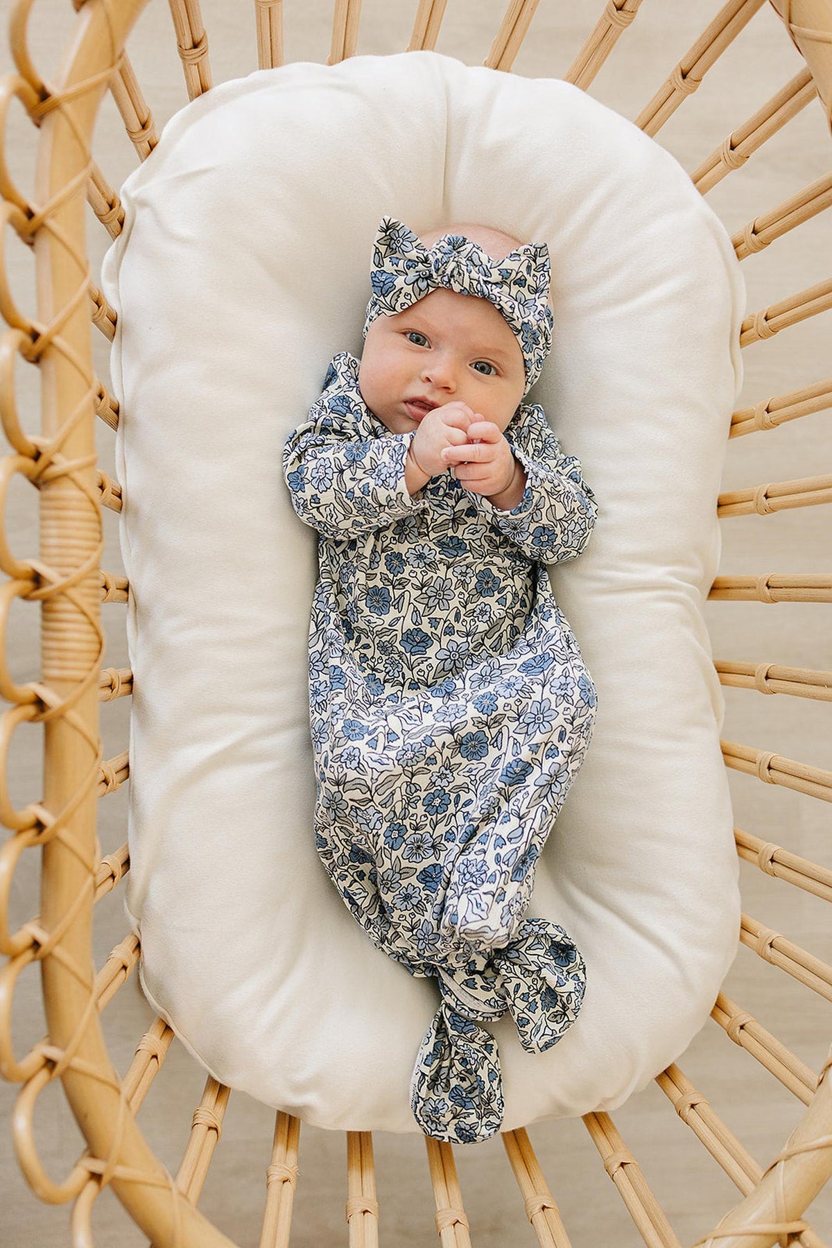 A baby in a floral-patterned gown and matching headband lies in a cushioned wicker bassinet.