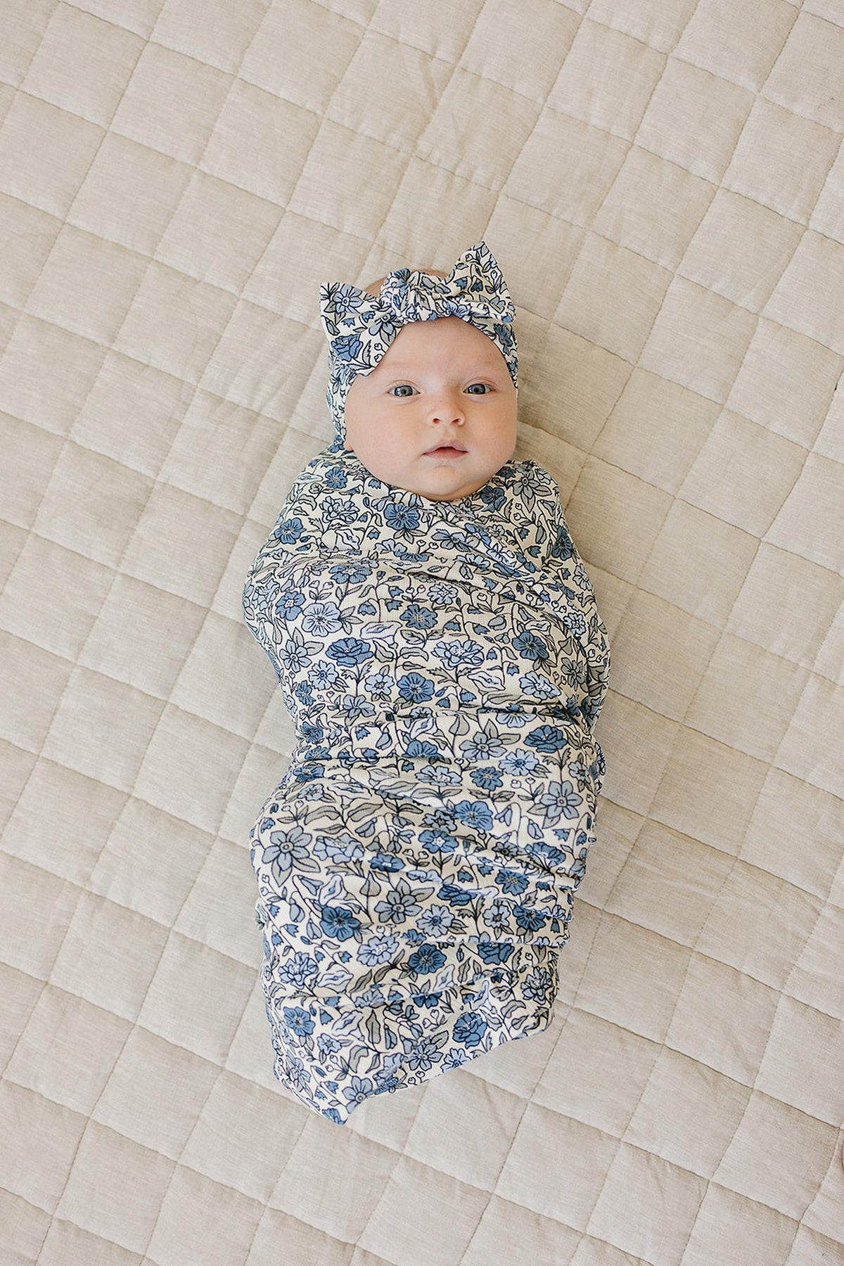 Baby wrapped in a blue floral-patterned swaddle and matching head wrap, lying on a beige quilted blanket.