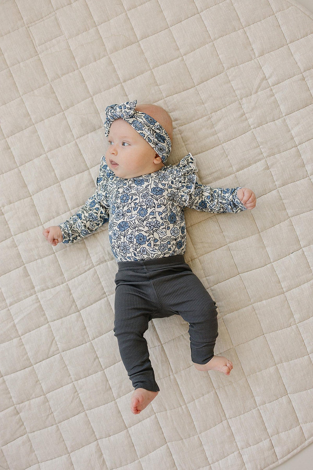 Baby wearing a floral ruffle sleeve bodysuit and headband, lying on a quilted beige blanket.