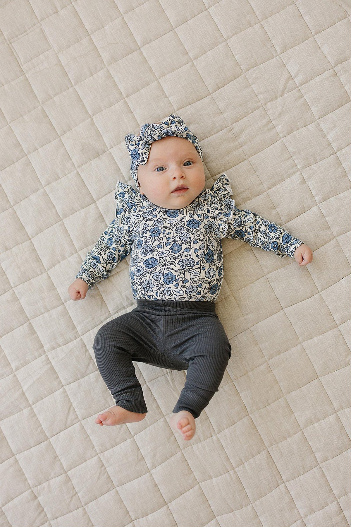 A baby lies on a quilt wearing a floral ruffle sleeve bodysuit with matching headband and dark pants.