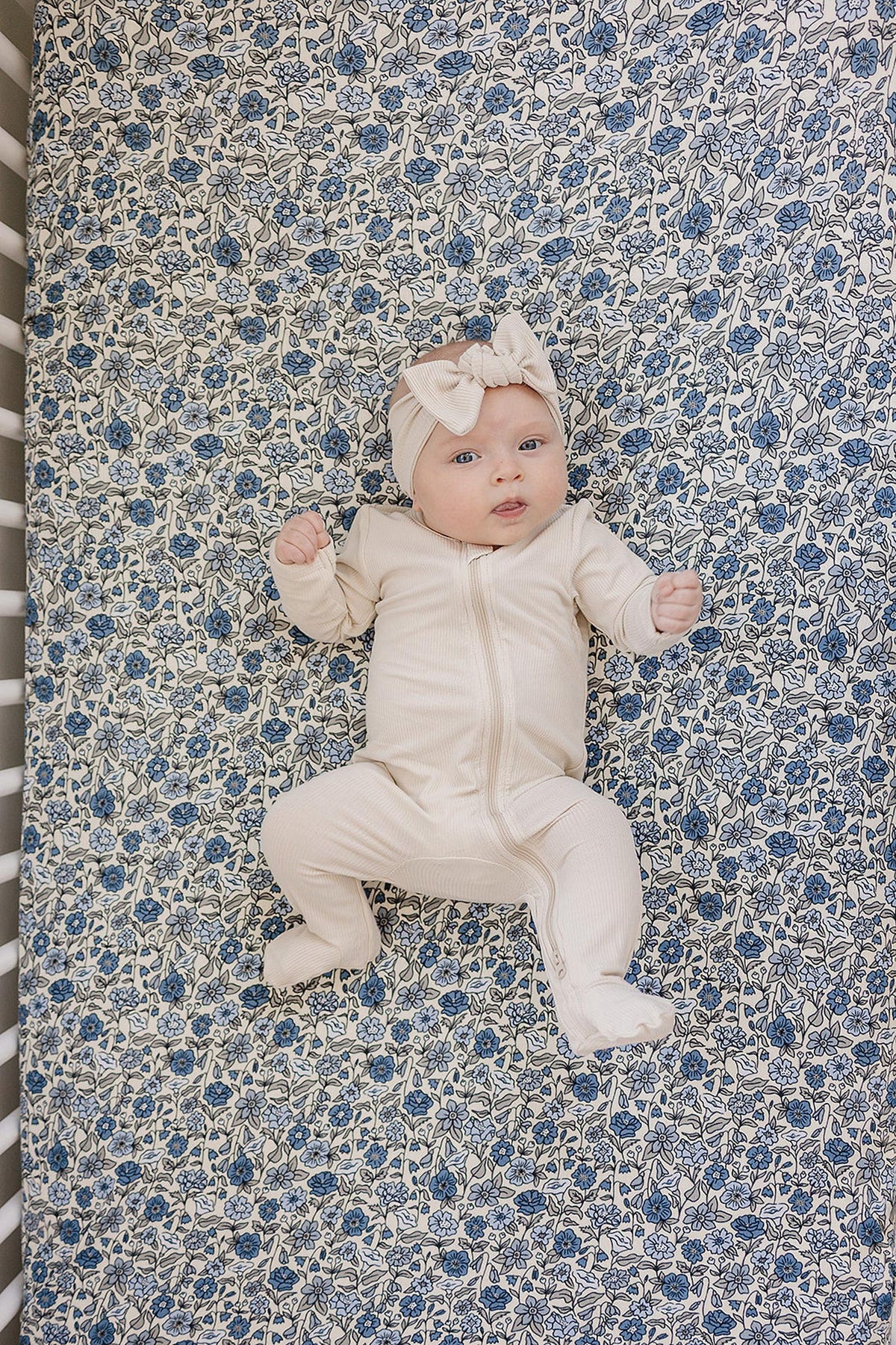 Baby in cream outfit and headband lying on a blue floral crib sheet.