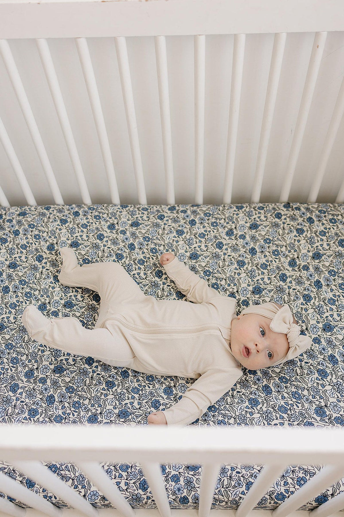 A baby in a cream outfit lies on a blue floral crib sheet in a white crib.