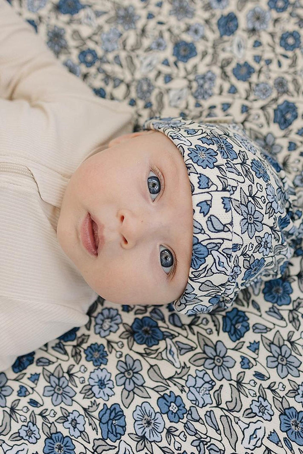 A baby wearing a floral-patterned hat and lying on a matching blanket with blue and white flowers.