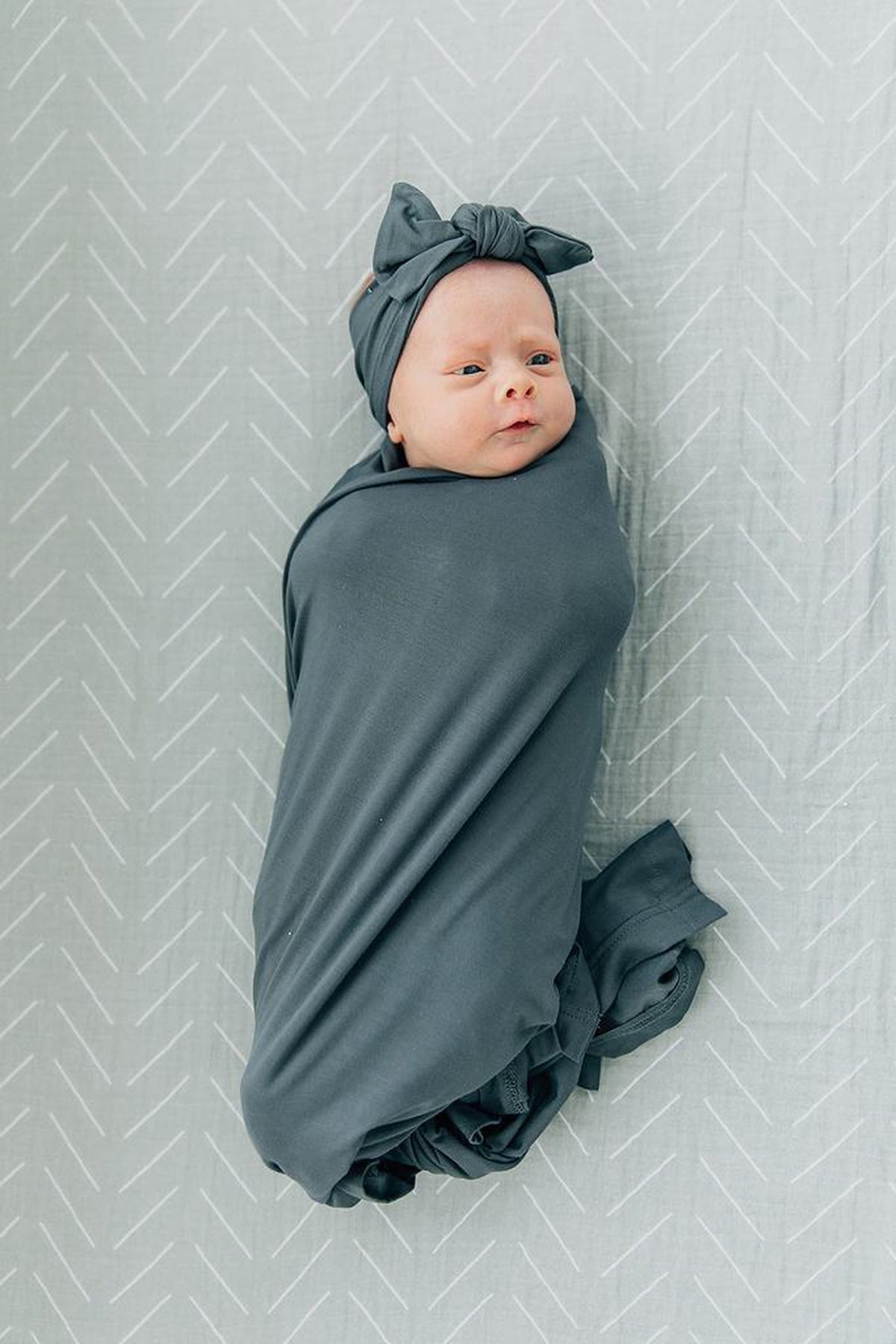 A baby wrapped in a charcoal swaddle with a matching head wrap lies on a patterned gray background.
