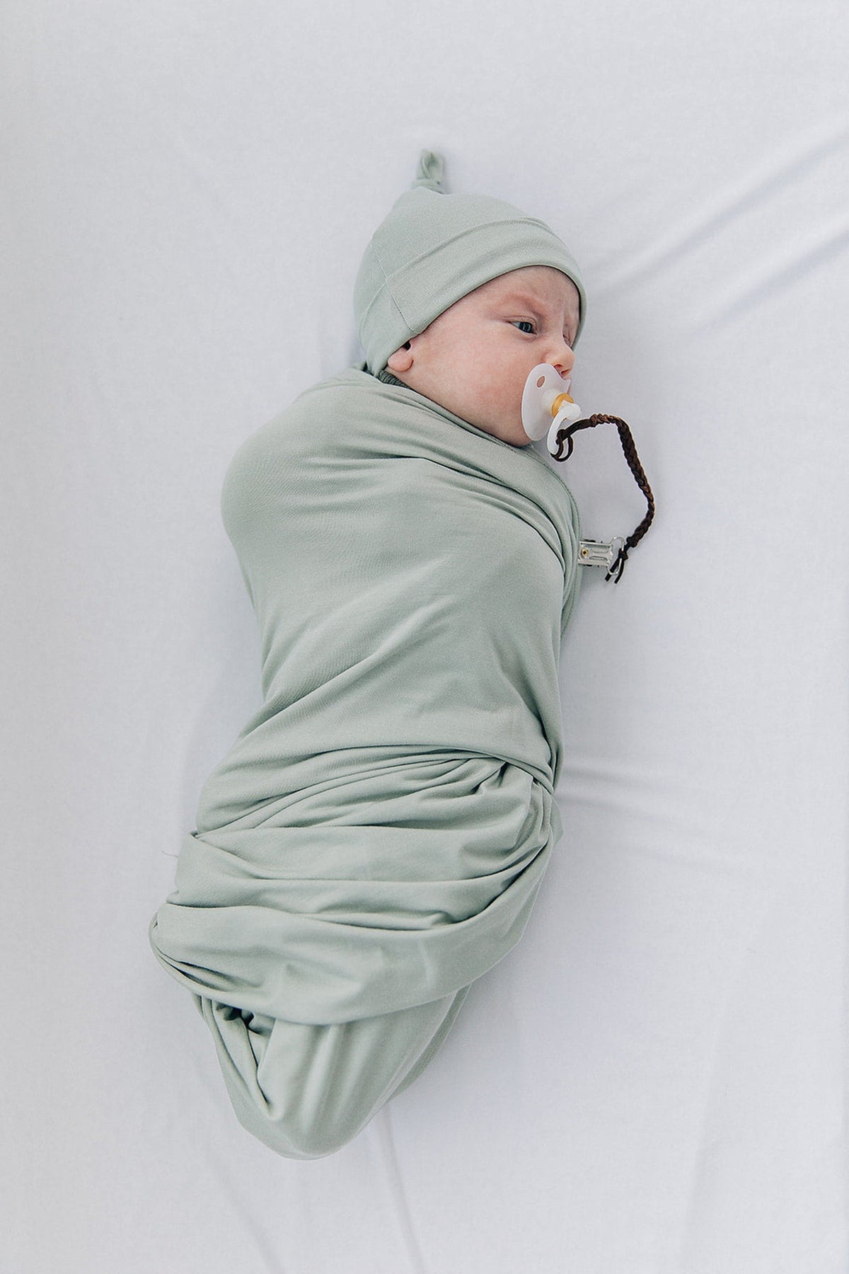 A newborn in a sage green bamboo swaddle and knot hat, holding a pacifier, lying on a white surface.