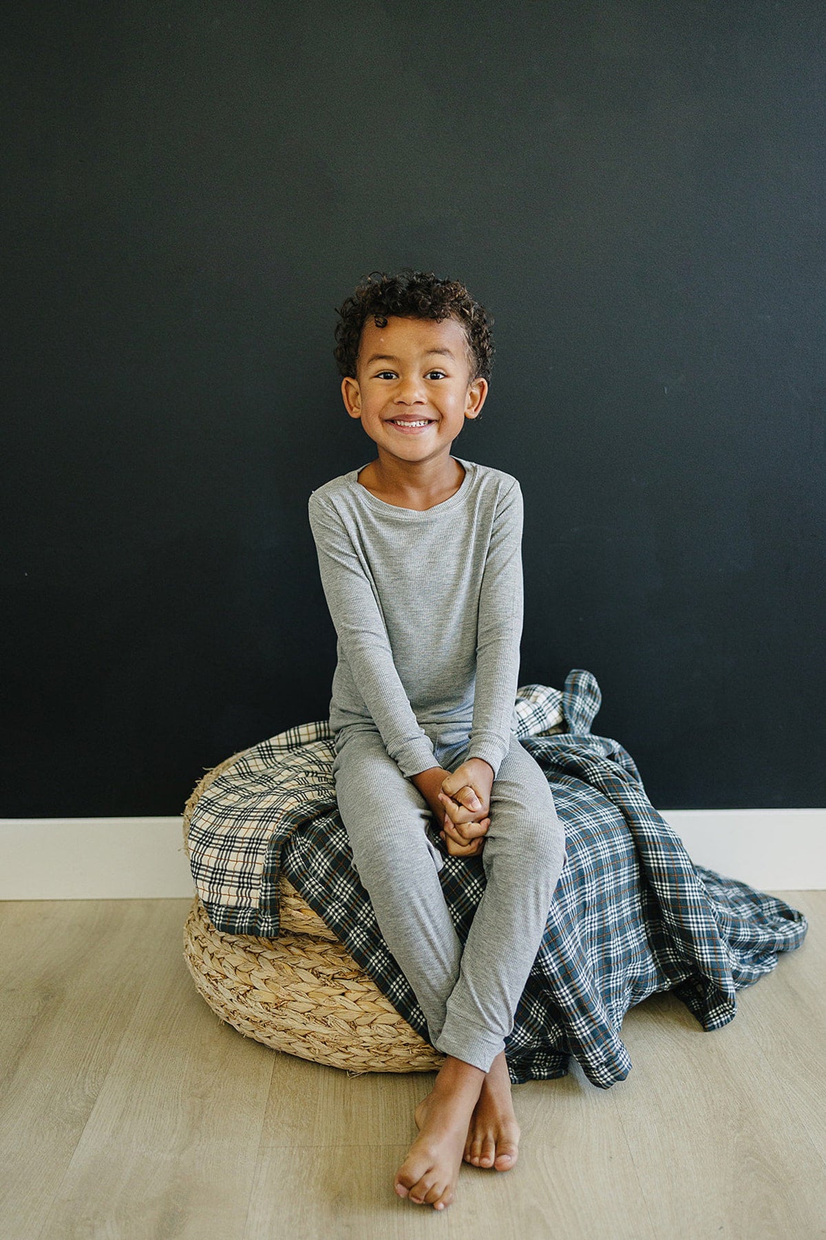 A child in gray pajamas sits on a wicker basket covered with a plaid quilt, smiling against a dark wall.