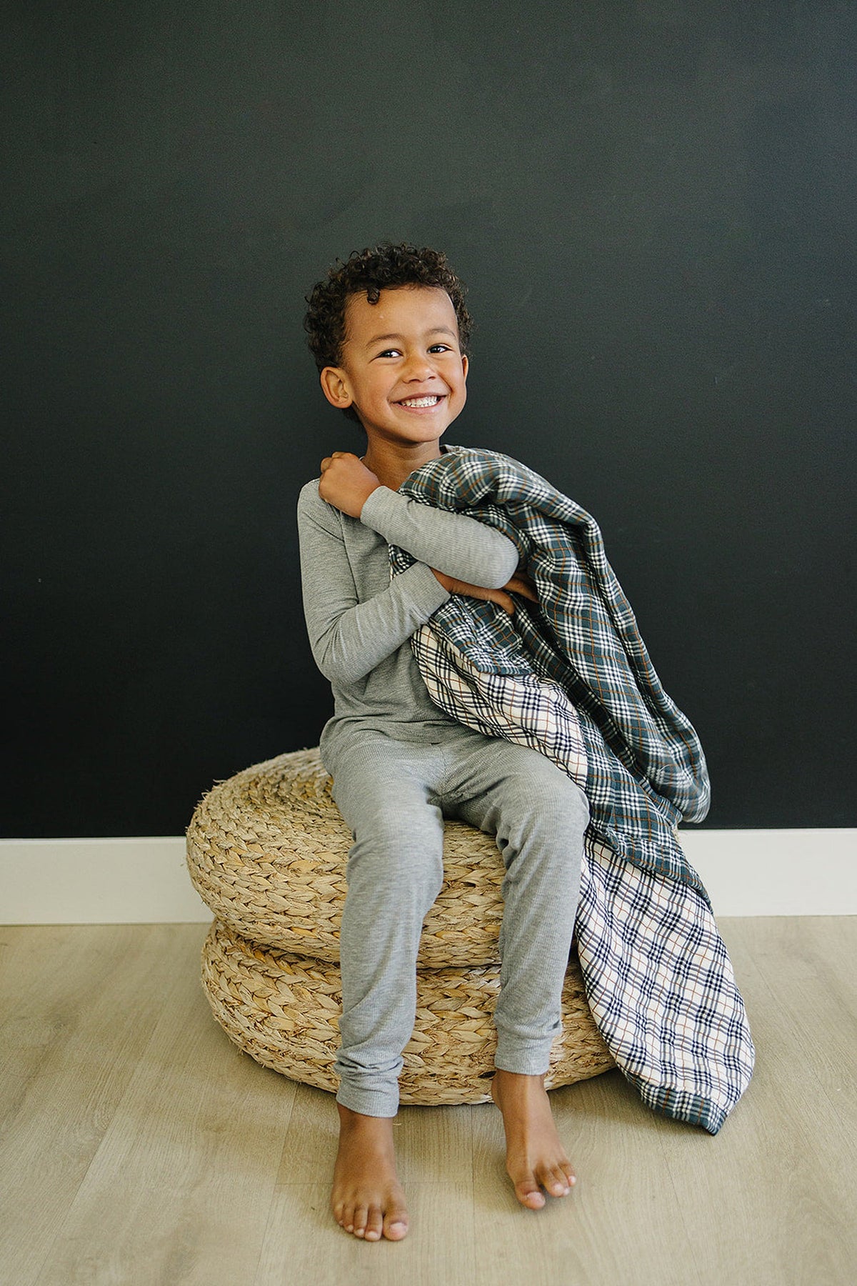 A smiling child in gray pajamas holds a plaid quilt while sitting on woven poufs against a dark wall.