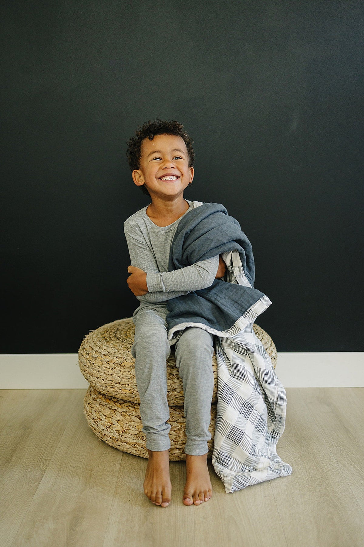 A smiling child in gray pajamas sits on a woven ottoman, holding a navy and plaid muslin quilt.