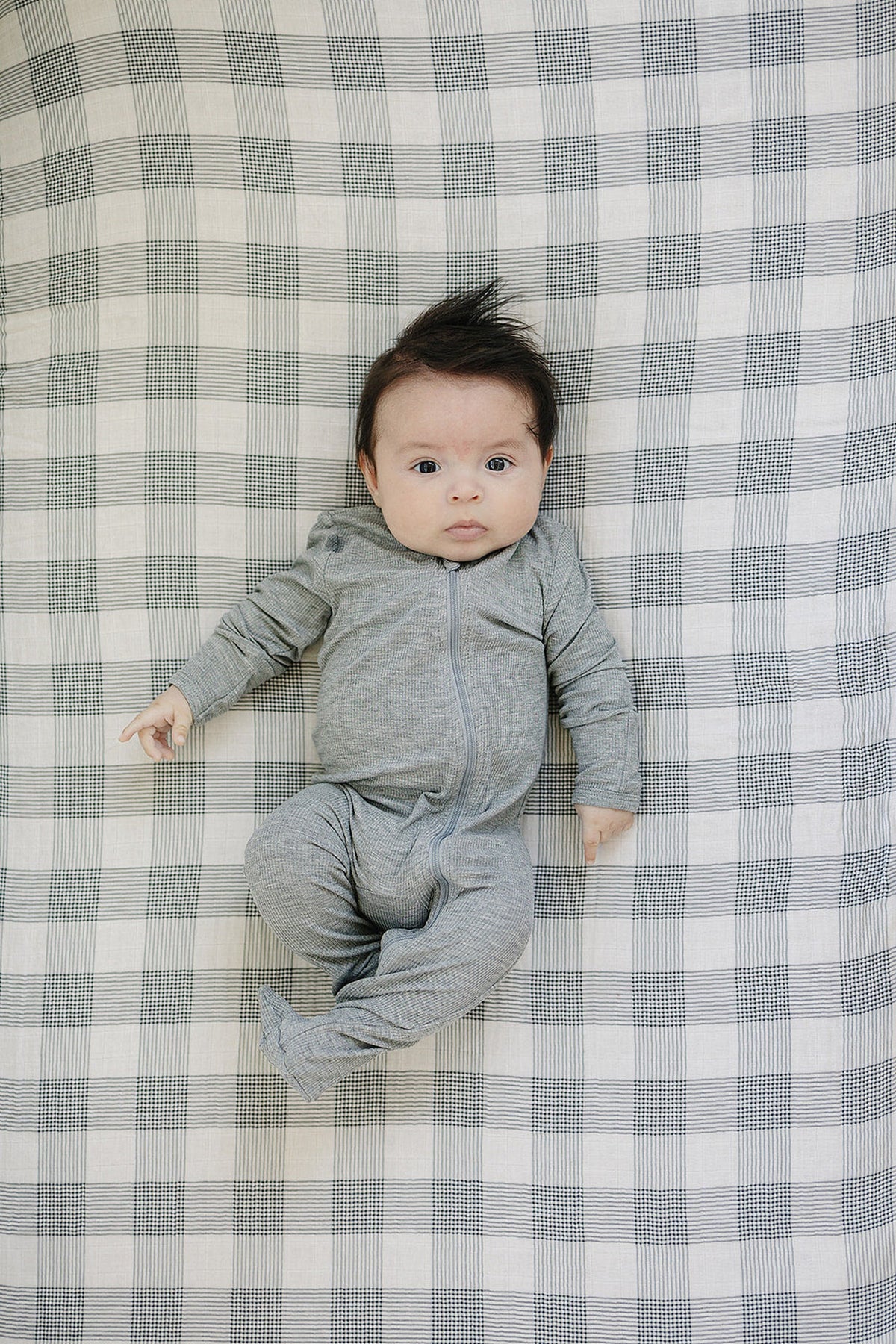A baby in a gray zip-up outfit lies on a coastal plaid muslin crib sheet with a soft, breathable texture.