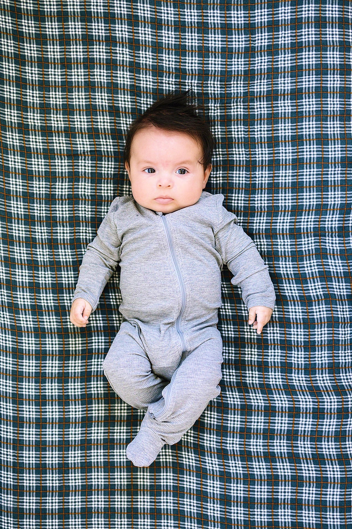 A baby in a gray onesie lies on a navy plaid crib sheet with a checkered pattern.