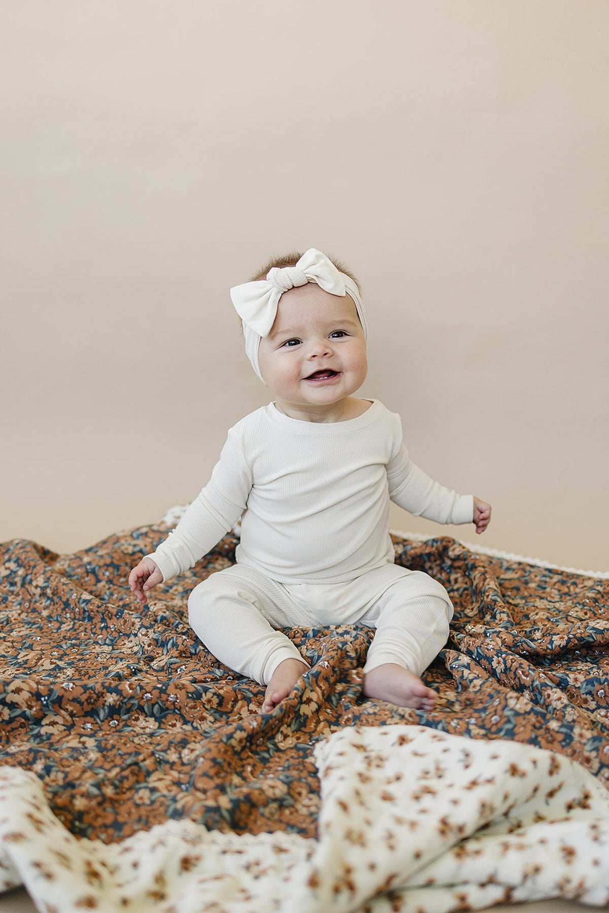 A smiling baby in white clothing sits on a floral-patterned quilt, wearing a matching headband.