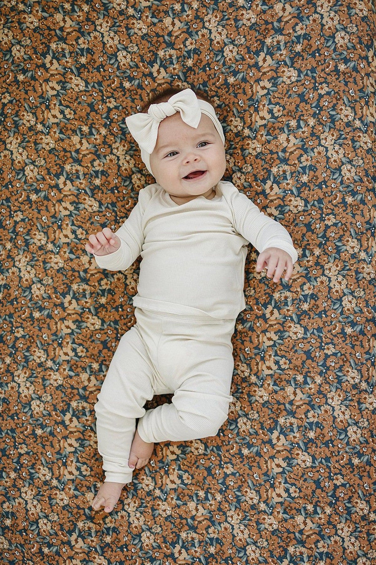 A smiling baby in cream clothing and bow headband lies on a floral-patterned crib sheet.