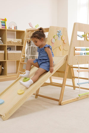 A young child in a blue romper slides down a wooden ramp of a multi-functional indoor climber, surrounded by colorful play accessories in a bright, airy playroom.