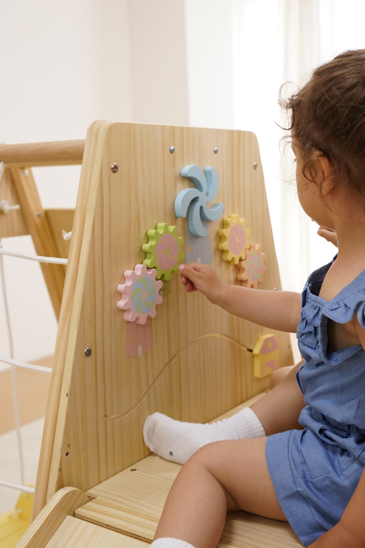 A young child interacts with colorful wooden gears on a light wood play climber. The indoor playset features a smooth finish, promoting safe and imaginative play for toddlers.