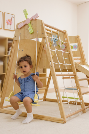 A young child sits on a swing attached to a wooden climbing frame, featuring natural wood and colorful accessories. The indoor playset encourages active play and imaginative movement for toddlers.