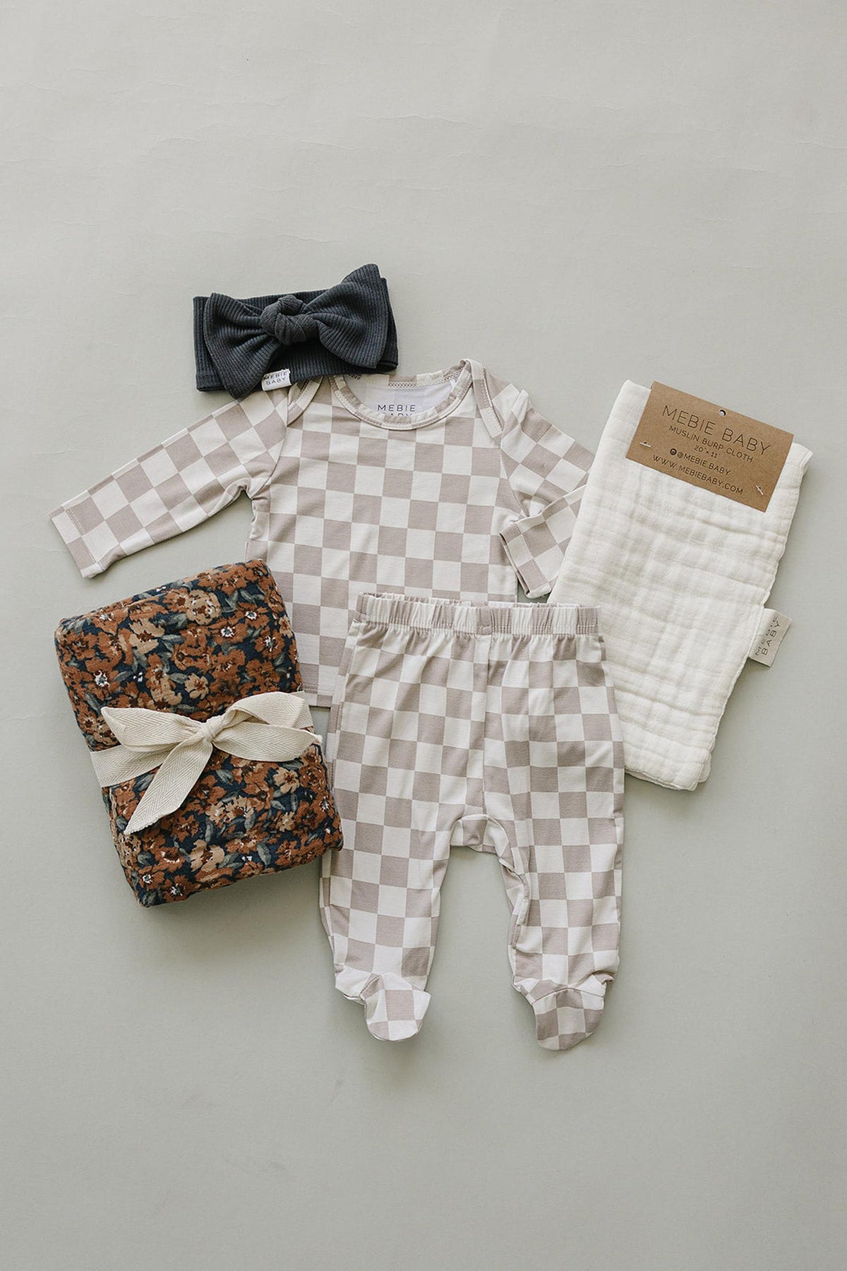 A taupe checkered bamboo layette set from Mebie Baby includes a long-sleeve onesie, pants, a floral blanket, a muslin cloth, and a dark headband, all arranged on a light background.