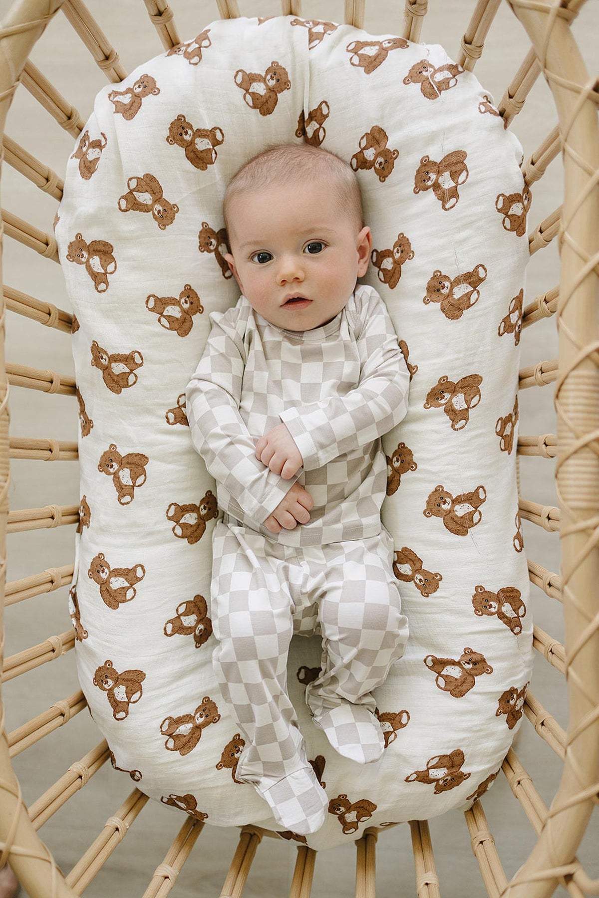 A baby lies in a woven bassinet, wearing a taupe checkered outfit. The soft, breathable fabric enhances comfort, while the bassinets padding features a playful bear print.