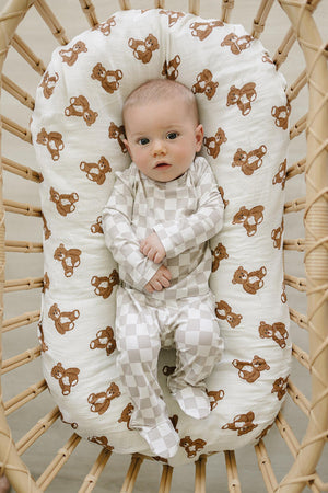 A baby lies in a woven bassinet, wearing a taupe checkered outfit. The soft, breathable fabric enhances comfort, while the bassinets padding features a playful bear print.