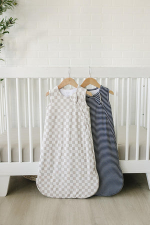 Two baby sleep sacks hang on wooden hangers against a white brick wall. One features a beige checkered pattern, while the other showcases a classic navy stripe design, both made.