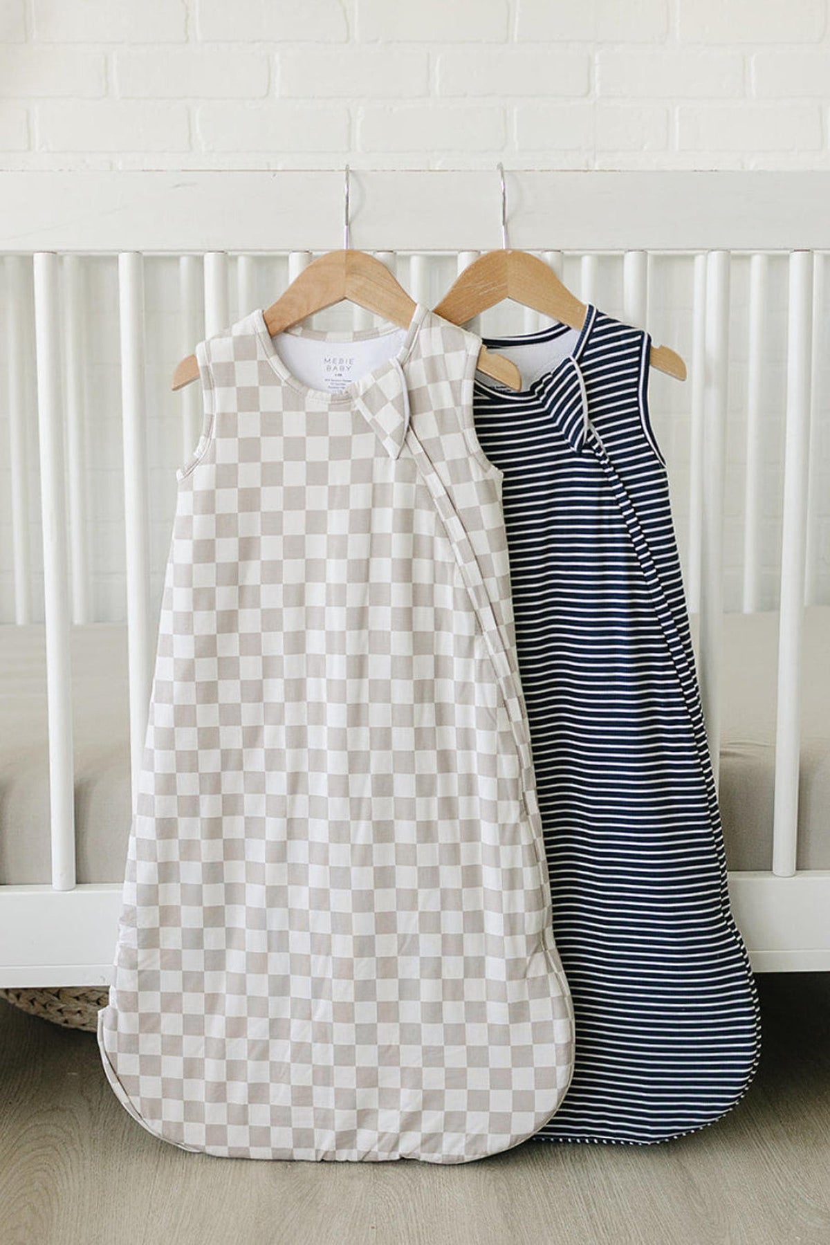 Two baby sleep sacks hang on wooden hangers against a white brick wall. One features a taupe checkered pattern, while the other has a navy and white striped design.