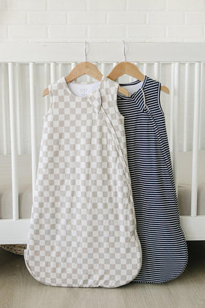 Two baby sleep sacks hang on wooden hangers against a white brick wall. One features a taupe checkered pattern, while the other has a navy and white striped design.