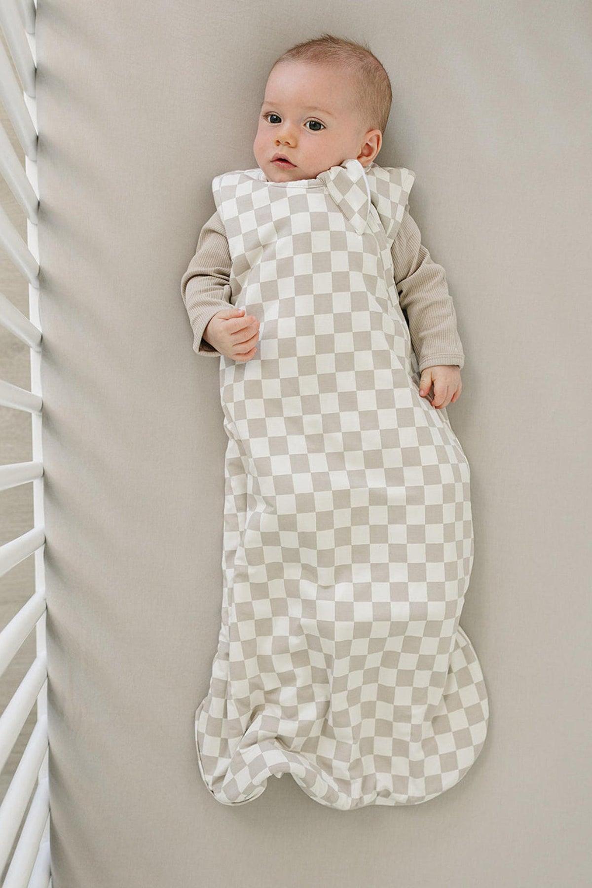 A baby lies peacefully in a taupe checkered sleep sack, made from soft, breathable fabric, on a neutral-toned crib mattress. The cozy design is perfect for keeping little ones comfortable.