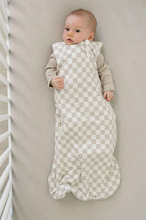 A baby lies peacefully in a taupe checkered sleep sack, made from soft, breathable fabric, on a neutral-toned crib mattress. The cozy design is perfect for keeping little ones comfortable.
