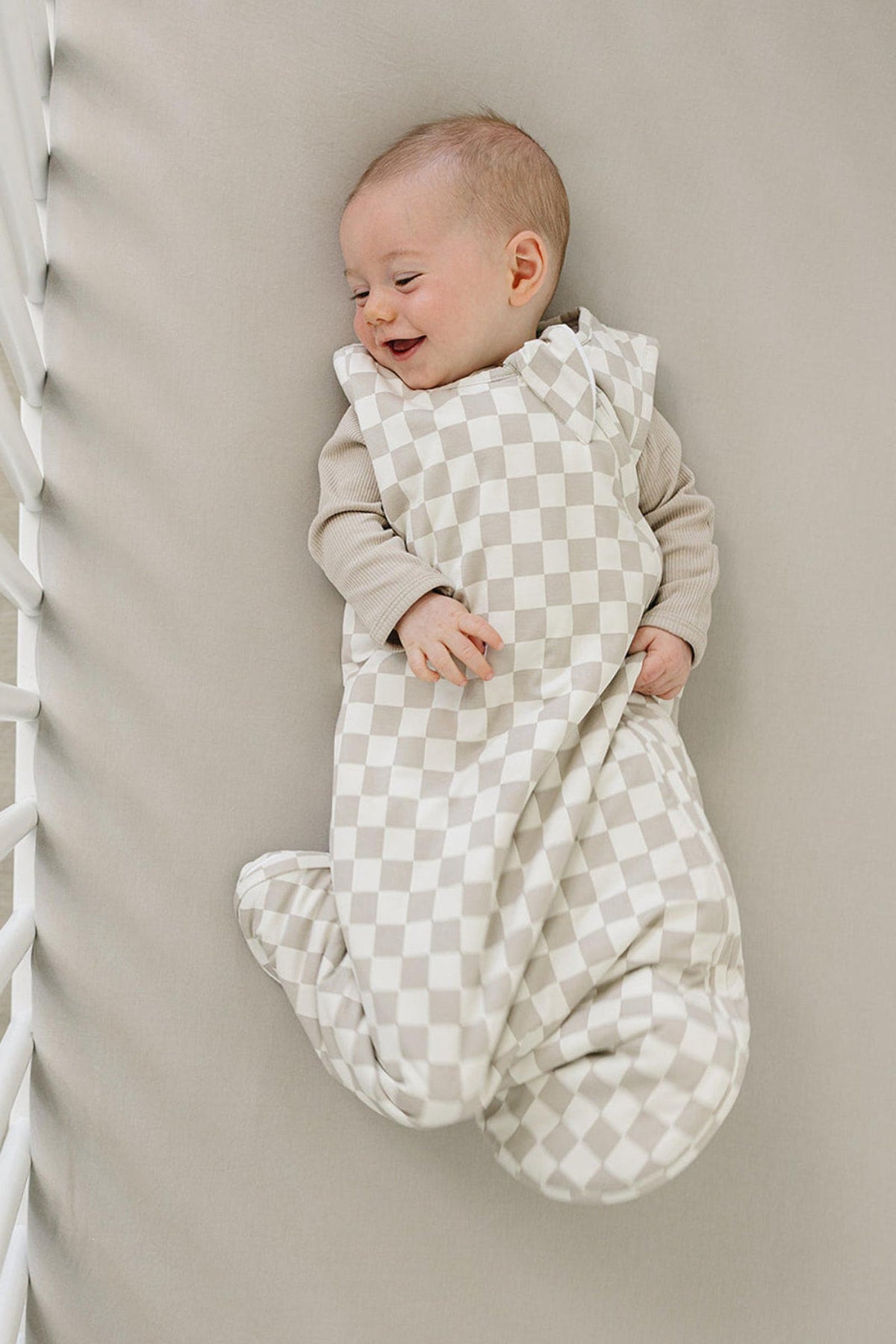A happy baby lies in a crib, wrapped in a soft taupe checkered sleep sack. The gentle fabric provides comfort.