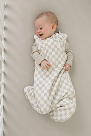 A happy baby lies in a crib, wrapped in a soft taupe checkered sleep sack. The gentle fabric provides comfort.