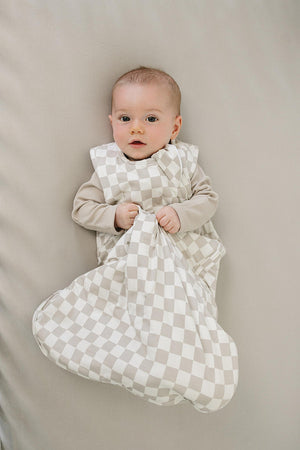 A baby lies on a soft, neutral-colored surface, wearing a taupe checkered bamboo sleep sack. The infant looks up, holding the sack, showcasing its cozy and breathable design.