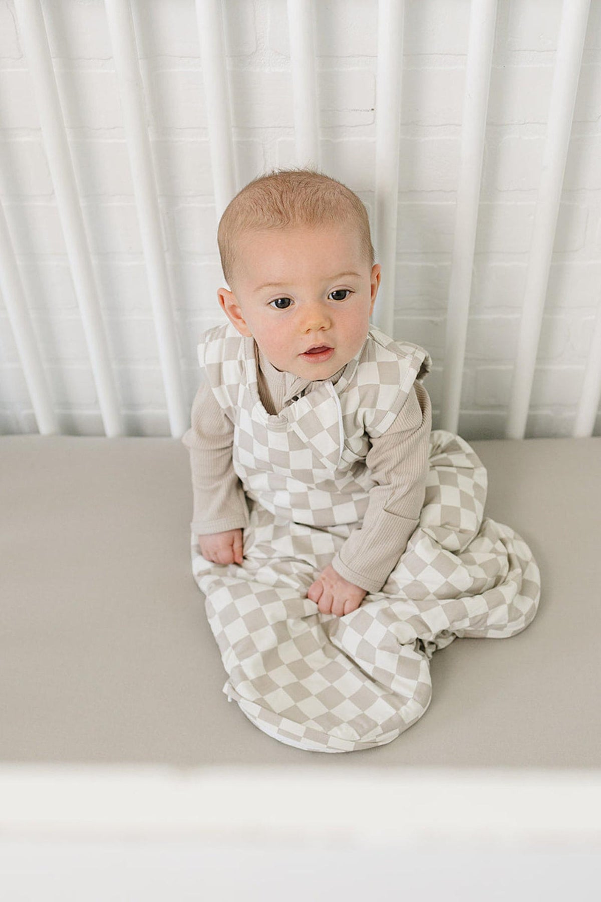 A baby sits in a crib wearing a taupe checkered bamboo sleep sack, featuring soft fabric and a cozy design.