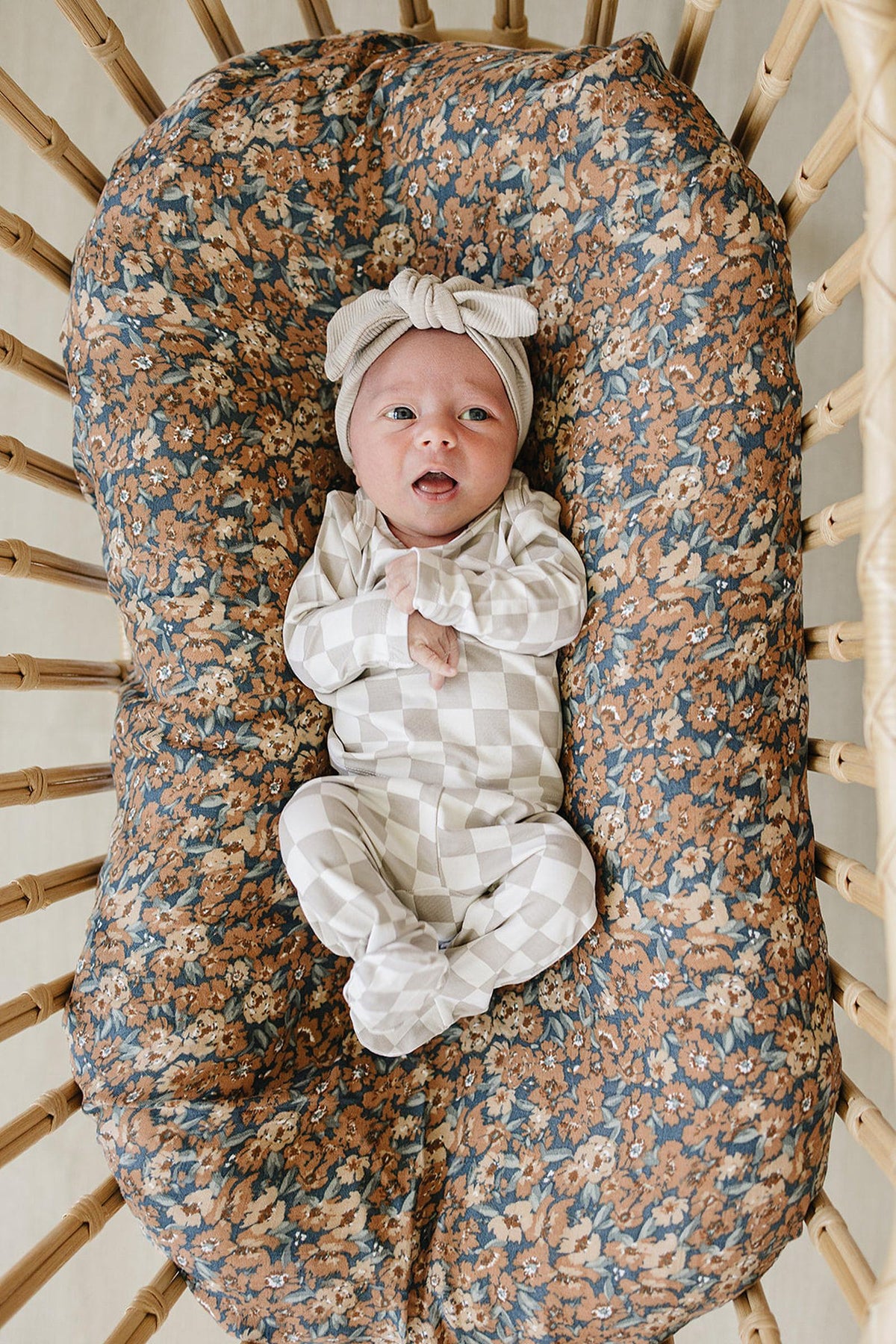 A baby lies comfortably in a woven bassinet, wearing a taupe checkered bamboo layette set. The bassinet is lined with a floral-patterned mattress cover, adding a cozy touch.