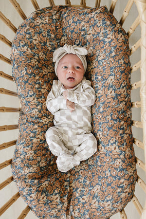 A baby lies comfortably in a woven bassinet, wearing a taupe checkered bamboo layette set. The bassinet is lined with a floral-patterned mattress cover, adding a cozy touch.