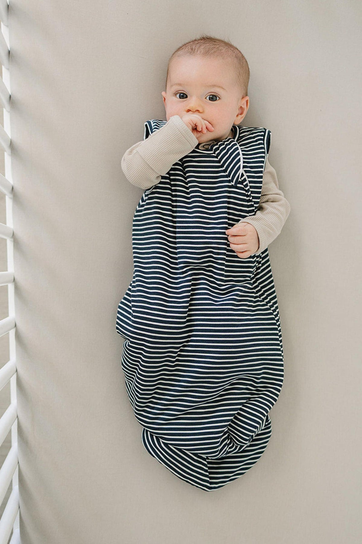 A baby lies comfortably in a navy stripe bamboo sleep sack, gently sucking their thumb. The soft, breathable fabric creates a cozy look against the neutral crib bedding.