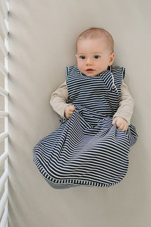 A baby comfortably rests in a crib, wearing a navy striped bamboo sleep sack. The soft fabric and classic stripes create a cozy, stylish look for peaceful sleep.