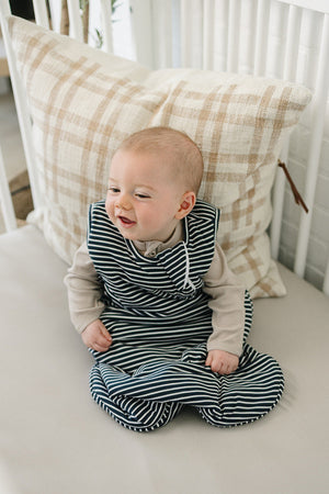 A smiling baby sits on a neutral-colored cushion, wearing a navy and white striped sleep sack. The soft fabric adds comfort, complementing the cozy nursery setting with a plaid pillow.