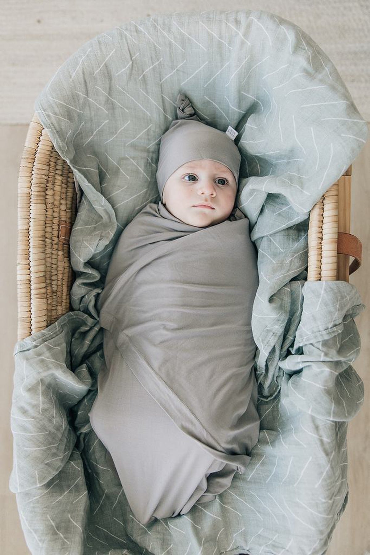 Newborn in a soft gray swaddle and knot hat, lying in a wicker basket with patterned fabric lining.